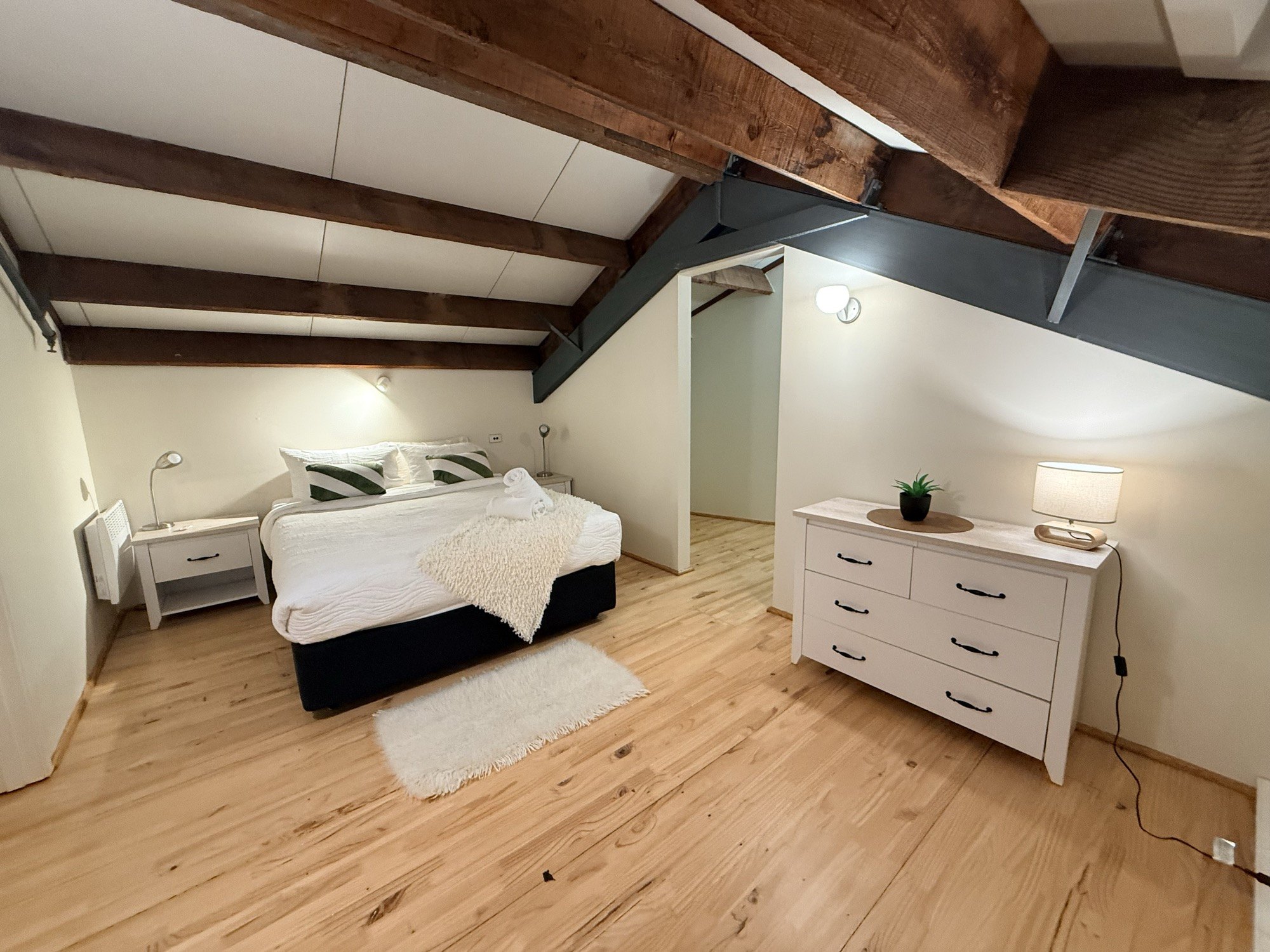 A beamed loft bedroom with white linen, soft lamplight and warm wood floors