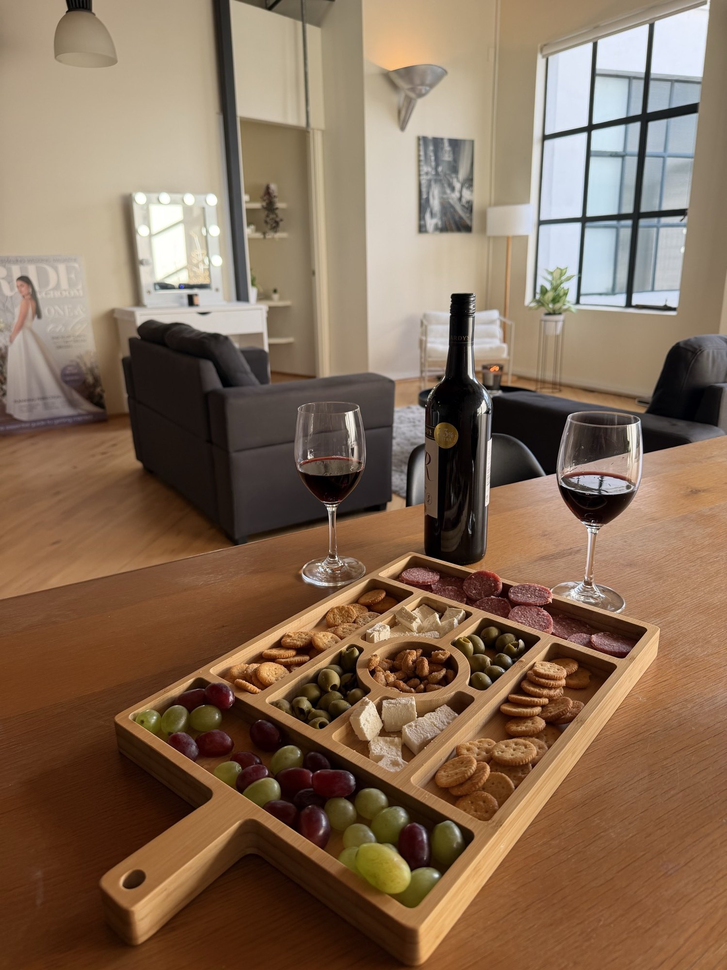 A grazing board with cheese and grapes, two glasses of red wine and a bottle on the loft table, the open living space and tall windows behind